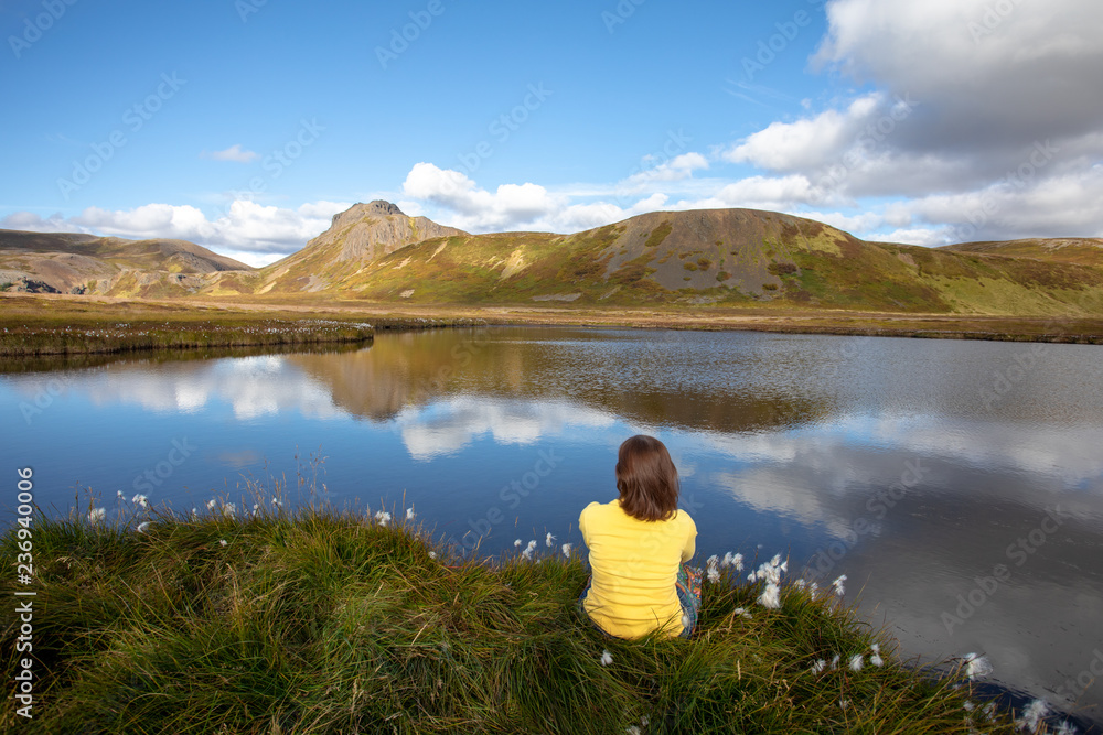 A young woman is sitting on the shore of a mountain lake. Clouds of mountains, and blue sky reflected in water. Summer view of travel. Iceland Europe