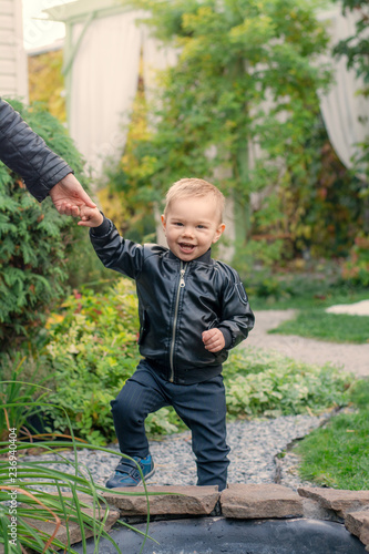  A little boy in the park holds up a hand. A child learns to walk. Fashionable boy laughs