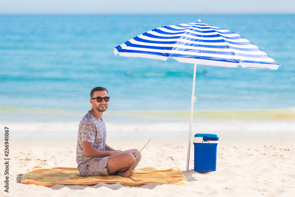 Young man relaxes on the beach under umbrella with laptop surf in internet.