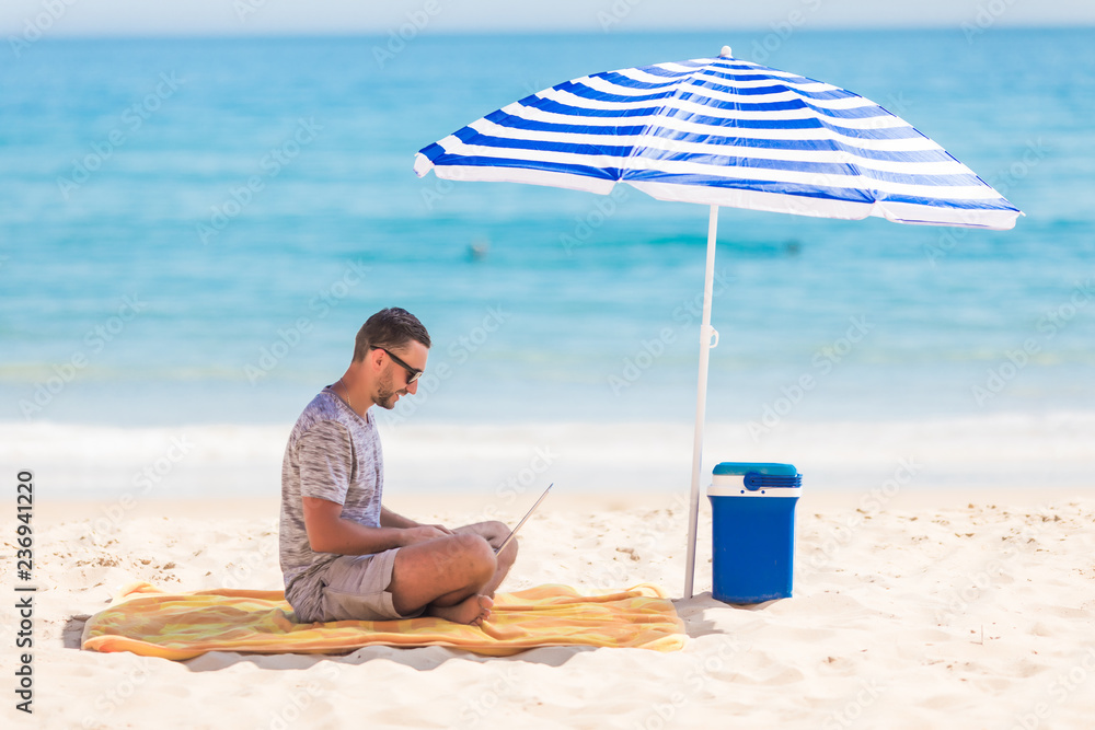 Young man with laptop working on the sand beach under umbrella on sea cost.