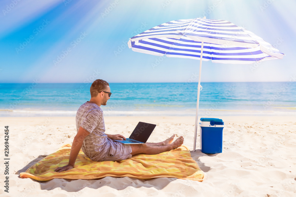 Young handsome man lusing laptop computer on beach bench with blue solar umbrella overhead, surrounded by turquoise sea and beautiful sky.