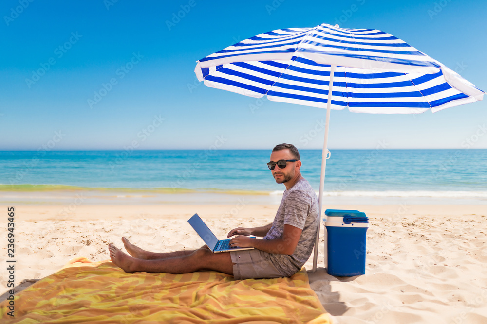 Young man using a laptop computer on the beach under solar umbrella. Freelance Concepts
