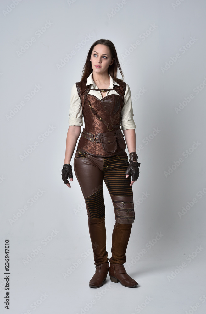 full length portrait of brunette girl wearing brown leather steampunk outfit. standing pose on grey studio background. full length portrait of brunette girl wearing brown leather steampunk outfit. standing pose on grey studio background.