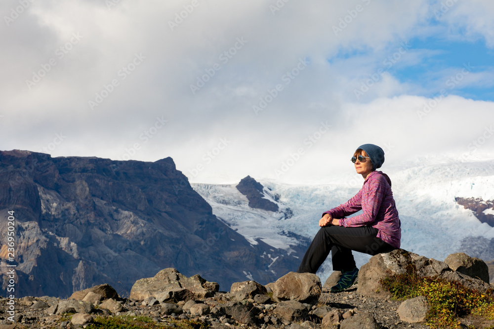 Fototapeta premium Young woman resting, sitting on the rocks in the mountains. Beautiful sunny view of the mountains and snow