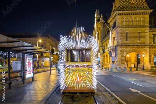 Budapest, Hungary - Festively decorated light tram (Fenyvillamos) on the move at Fovam Square with the Great Market Hall (Vasarcsarnok) by night. Christmas season in Budapest photo