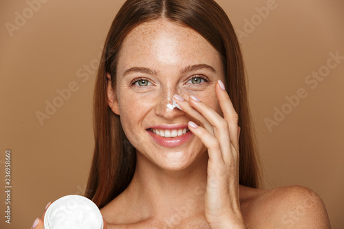 Beauty image of attractive shirtless woman smiling and holding jar with face cream, isolated over beige background photo