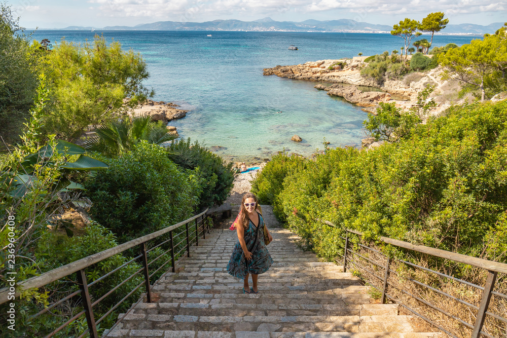 Tourist girl on the beach on a wonderful stairway leading to the sea with clear turquoise waters in the background with sunglasses dress and handbag on the island of Mallorca, Spain.