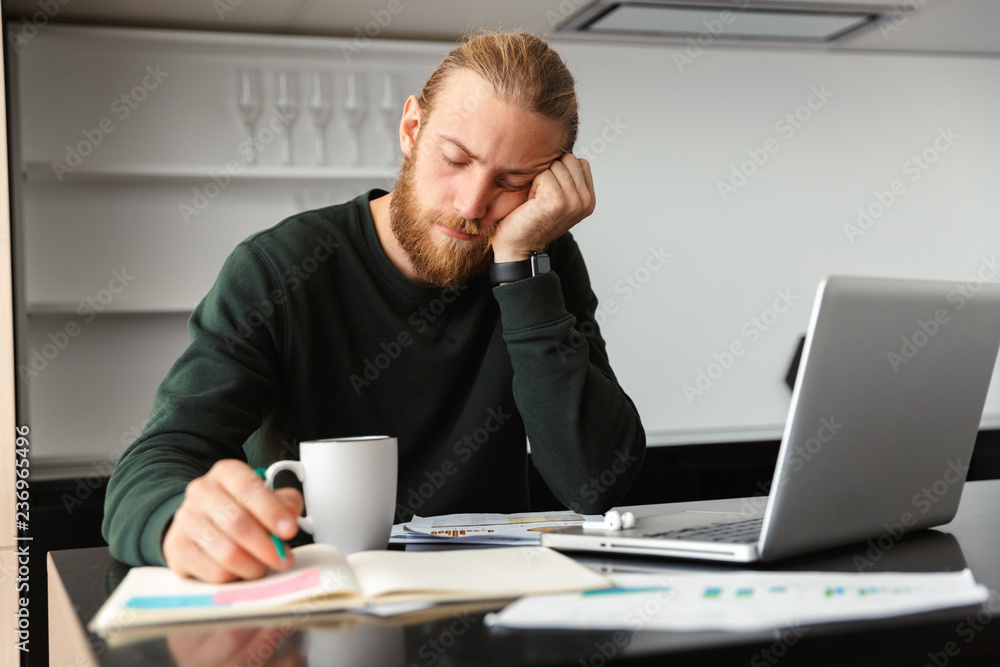 Tired young bearded man sitting at the kitchen drinking coffee using laptop computer. Tired young bearded man sitting at the kitchen drinking coffee using laptop computer.