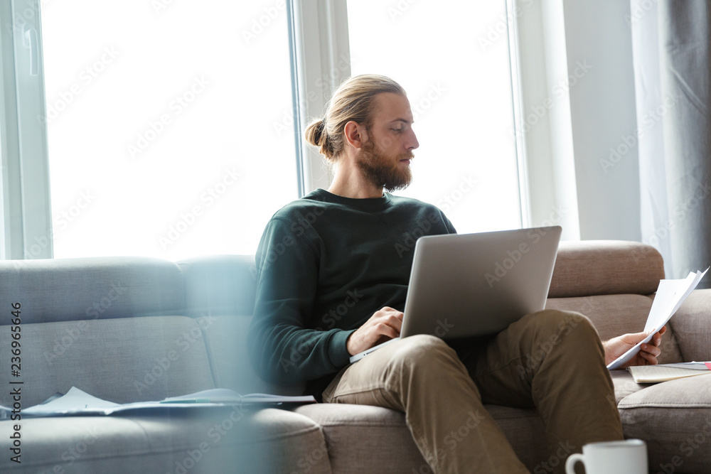 Concentrated young bearded man sitting in home using laptop computer. Concentrated young bearded man sitting in home using laptop computer.