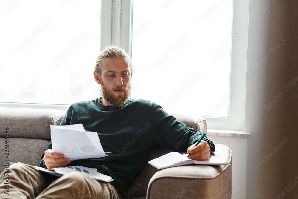 Concentrated young bearded man sitting in home work with documents.