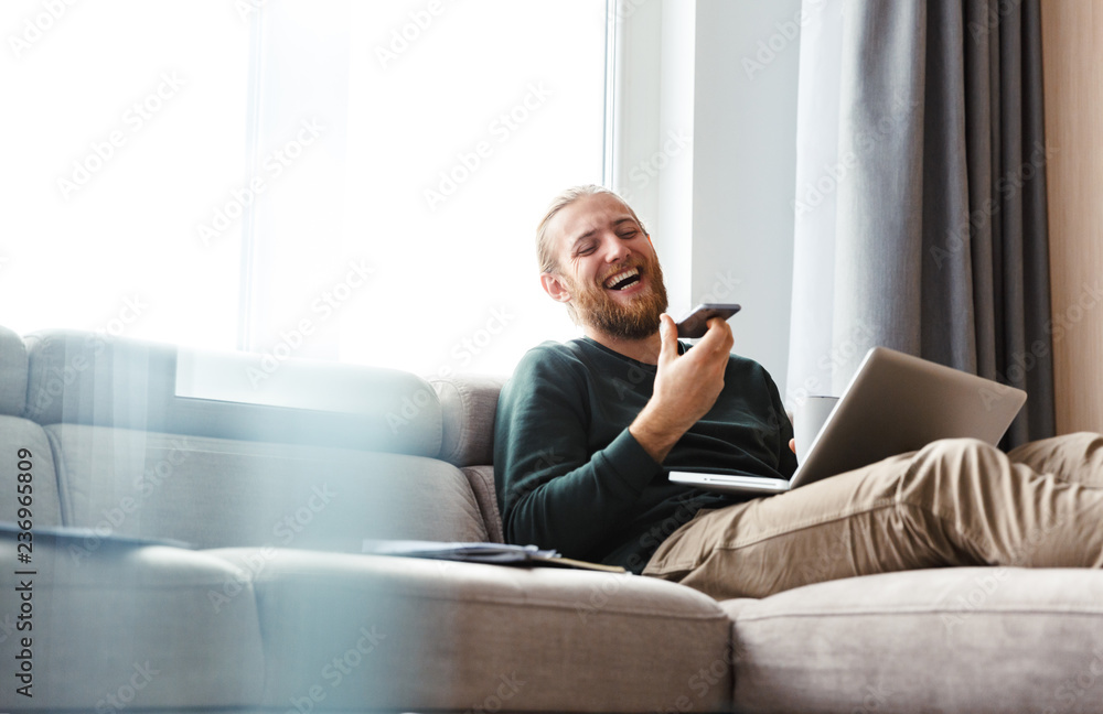 Fototapeta premium Young bearded man sitting in home using laptop computer and mobile phone.