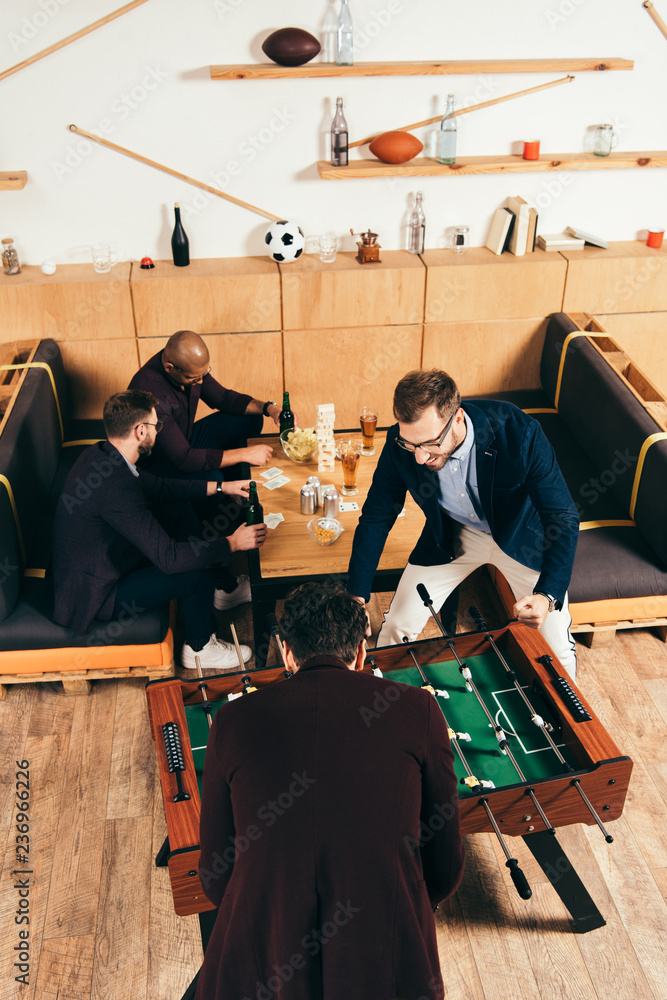 high angle view of businessmen playing table football while multiethnic colleagues resting on sofa in cafe