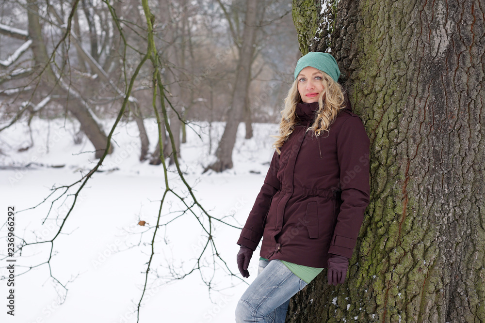 blond woman leaning against tree in snow covered park in winter blond woman leaning against tree in snow covered park in winter