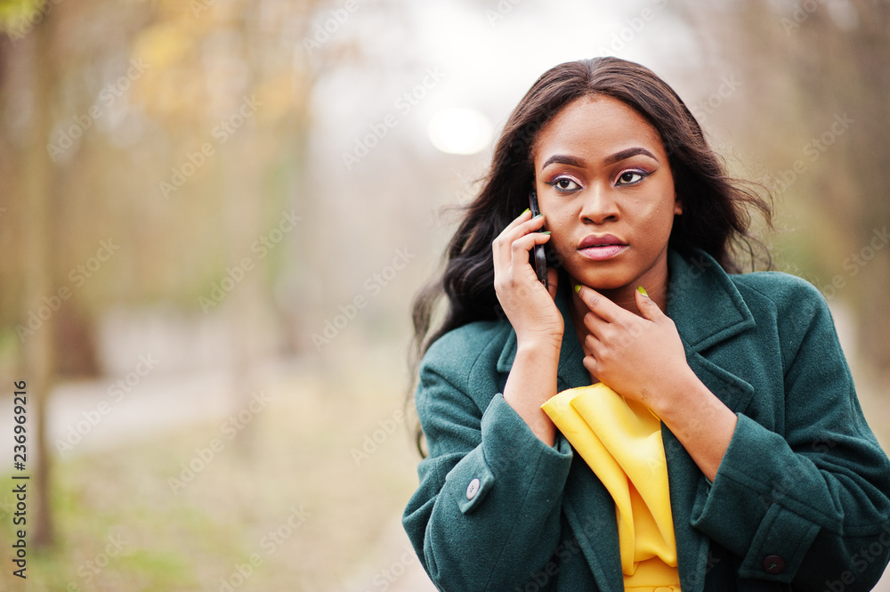 Stylish african american woman at green coat and yellow dress posed against autumn park and speaking on phone. Stylish african american woman at green coat and yellow dress posed against autumn park and speaking on phone.
