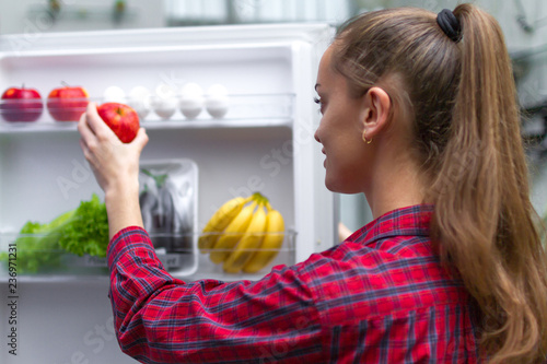 Young, attractive girl pulls out a fresh apple for a snack from the fridge. Food storage, vegetables and fruits in the refrigerator. Proper, healthy and fresh food. photo