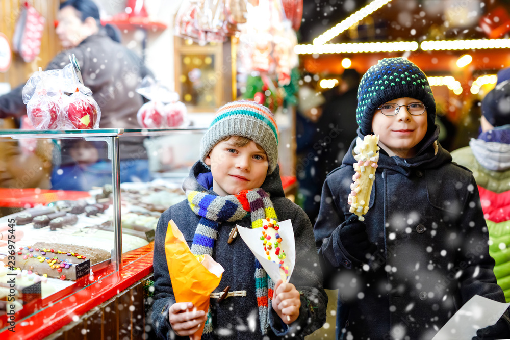 Two little kid boys, cute siblings eating bananas covered with chocolate, marshmellows and colorful sprinkles near sweet stand with gingerbread and nuts. Happy children on Christmas market in Germany.