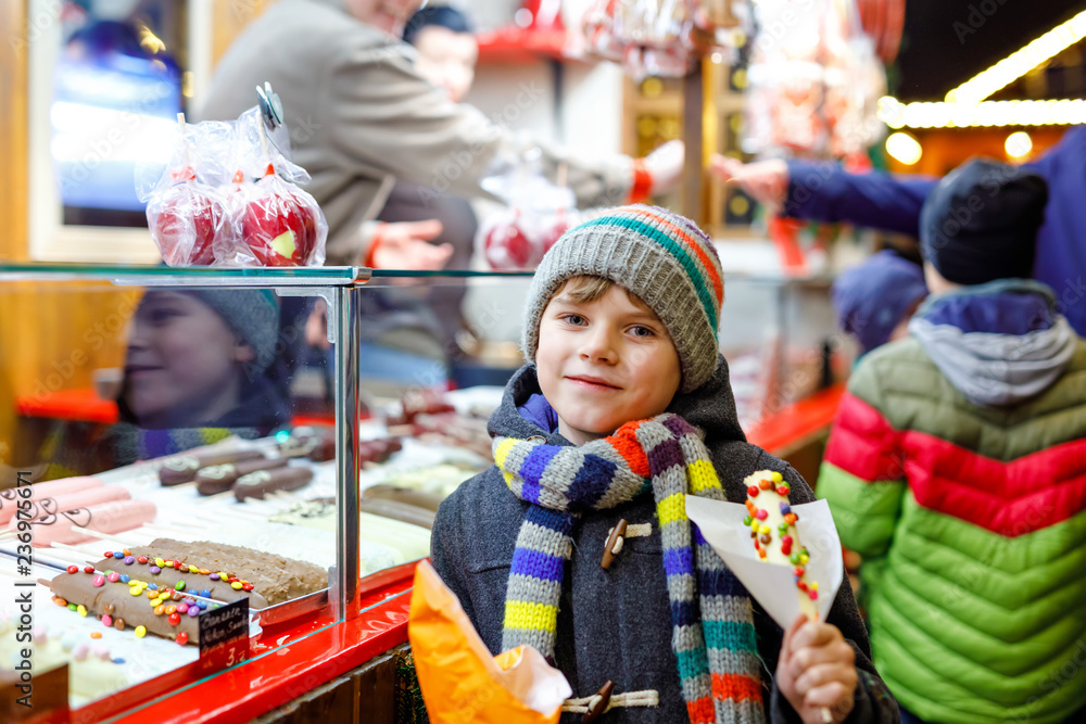 Little kid boy, cute child eating bananas covered with chocolate, marshmellows and colorful sprinkles near sweet stand with gingerbread and nuts. Happy boy on Christmas market in Germany.