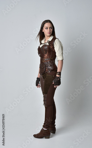 full length portrait of brunette girl wearing brown leather steampunk outfit. standing pose on grey studio background.