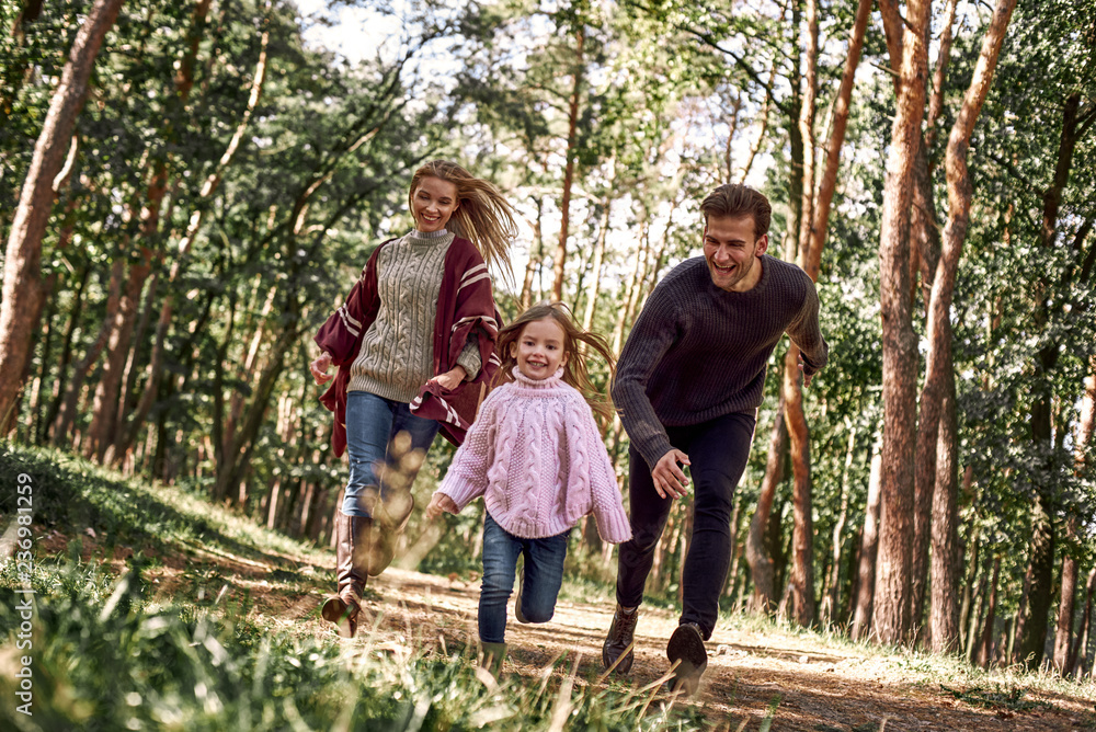 Happy parents with their daughter hold hands and run at forest path