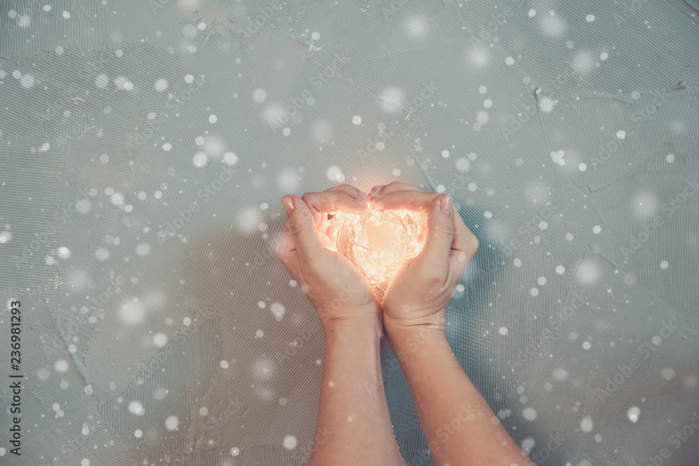 woman's hand making shape of a heart, holding warm Christmas lights, wooden background, top view woman's hand making shape of a heart, holding warm Christmas lights, wooden background, top view
