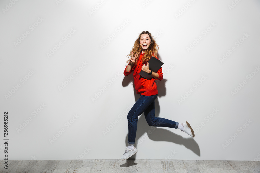 Horizontal image of beautiful woman 20s wearing red sweatshirt walking and holding gray laptop, isolated over white background