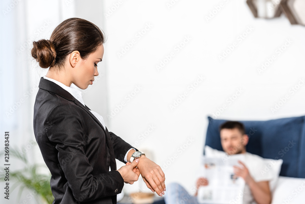 girlfriend in suit checking time at wristwatch and boyfriend in pajamas reading newspaper in bedroom in morning, gender equality concept girlfriend in suit checking time at wristwatch and boyfriend in pajamas reading newspaper in bedroom in morning, gender equality concept