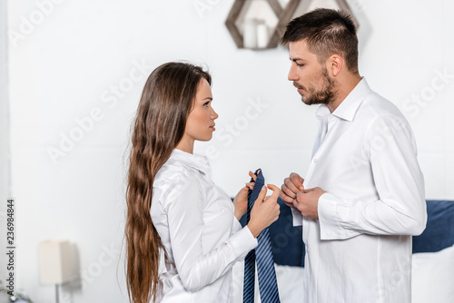 handsome boyfriend buttoning shirt, girlfriend holding his tie in weekday morning in bedroom, social role concept