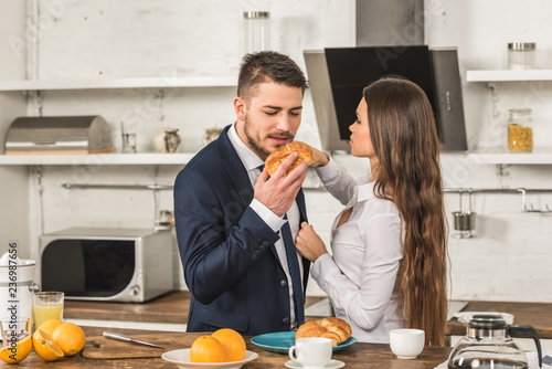 boyfriend eating croissant and girlfriend fixing his jacket in kitchen, sexism concept