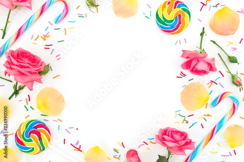 Round frame of roses flowers with pastel petals and bright sugar candies on white background. Flat lay, top view.