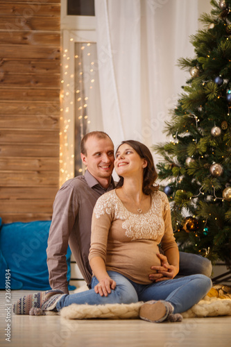 Photo of cheerful pregnant couple on background of decorated New Year tree,Cristmas interior