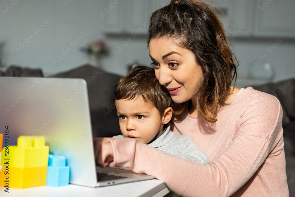 surprised little boy and his mother using laptop at table in living room at home surprised little boy and his mother using laptop at table in living room at home