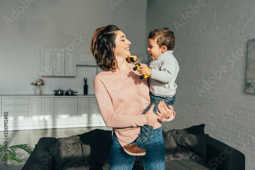 happy woman holding little boy with toy cars on hands at home