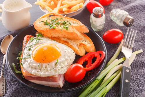 Fried egg with bacon in a black plate with fried pieces of bread, greens tomatoes, a jug of milk and French fries on a gray wooden table. Close-up
