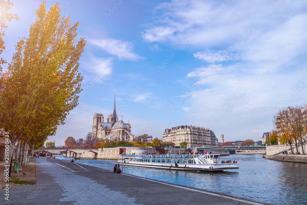 Obraz premium Cathedral of Notre Dame de Paris at sunny autumn afternoon. Embankment of the Seine. Boat on scenic route. Tourists a walk and relax in warm weather. Blurred unrecognizable faces. Paris. France.
