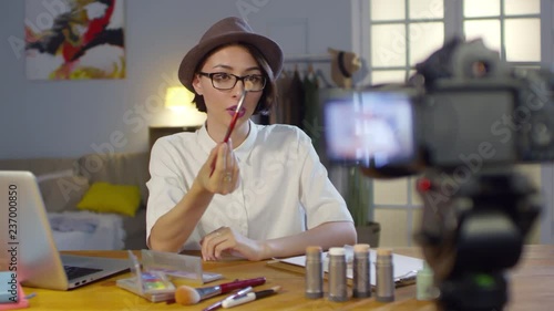 Medium shot of young woman in fedora hat and glasses recording makeup tutorial and explaining how to use products and brushes photo