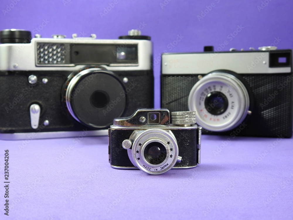 Photo cameras: in the foreground, the miniature of a vintage camera. Behind, two vintage blurred analog cameras. Lilac background.