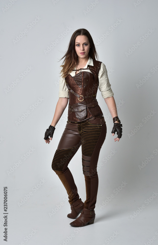 full length portrait of brunette girl wearing brown leather steampunk outfit. standing pose on grey studio background. full length portrait of brunette girl wearing brown leather steampunk outfit. standing pose on grey studio background.