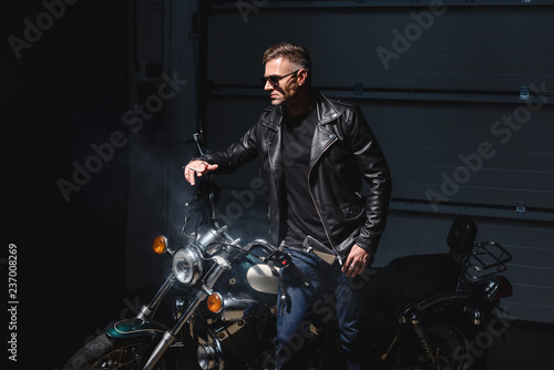 fashionable guy in black sunglasses standing by motorcycle in garage