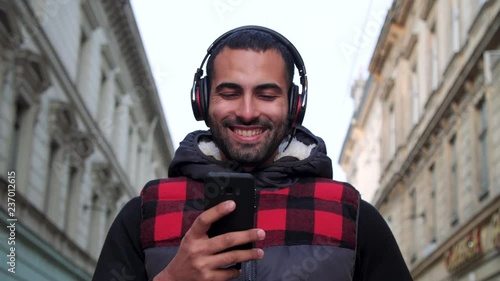 Dancing and Smiling Attractive Guy Using his Smartphone for Texting Massage. He’s Casual Dressed. Walking in Old European City Streets while Listening to Music. Headphones. Casual Lifestyle. People. photo