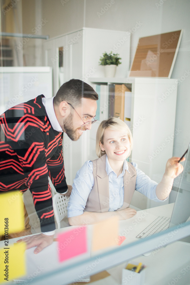 Fototapeta premium Positive excited young colleagues in casual outfits discussing marketing project in office: pretty lady pointing at monitor while explaining information to man