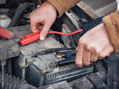 Man recharging a dead car battery in winter time using start-charger. Close up hands of a man with two contact clips from battery charger for vehicles. People under bonnet with starter cables © marmoset