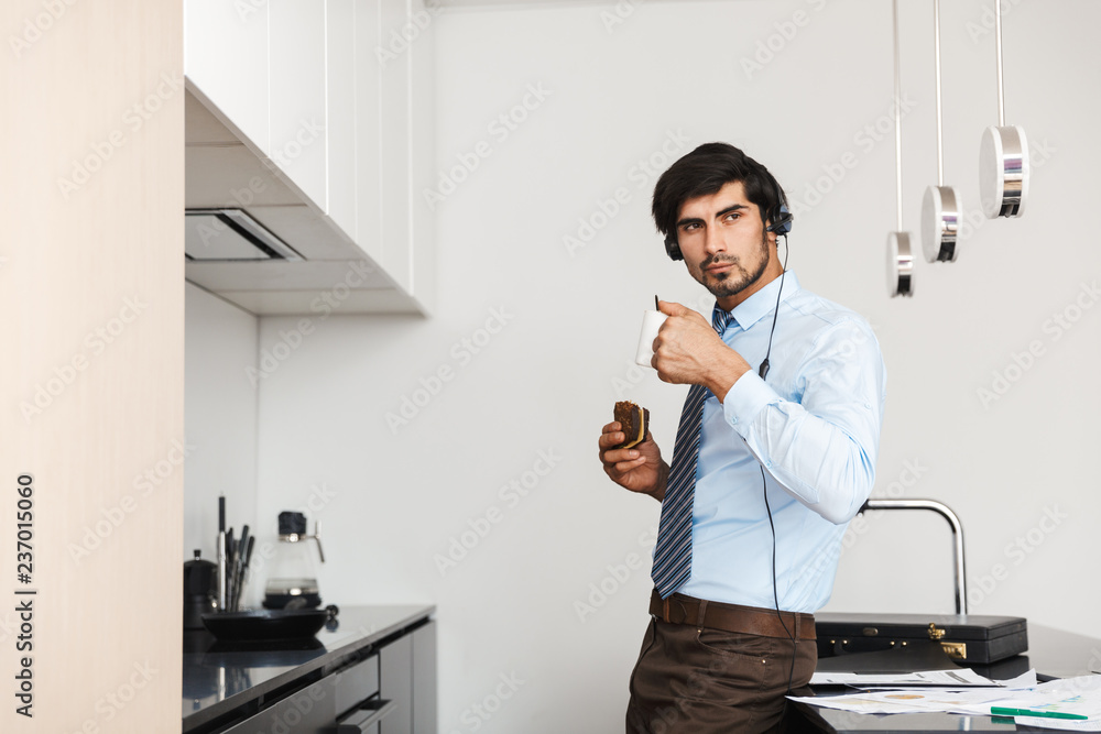 Business man at the kitchen eat sandwich using laptop computer wearing headphones drinking coffee. Business man at the kitchen eat sandwich using laptop computer wearing headphones drinking coffee.
