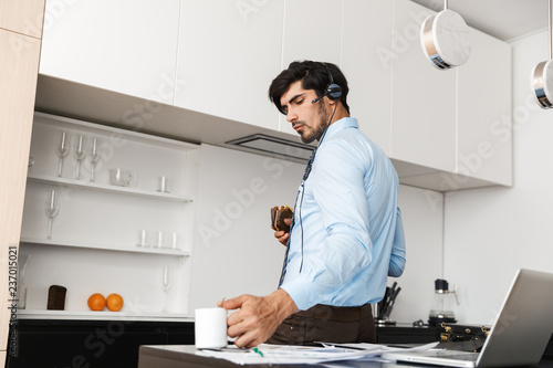 Business man at the kitchen eat sandwich using laptop computer wearing headphones drinking coffee.