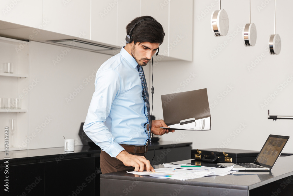 Concentrated young business man at the kitchen wearing headphones work with documents.