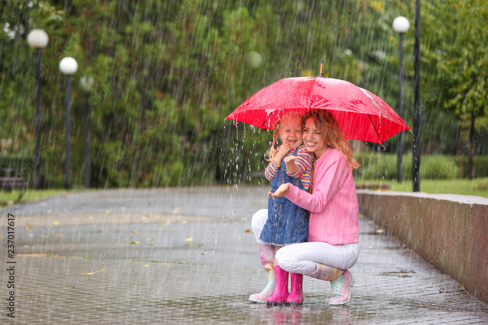 Naklejka premium Happy mother and daughter with red umbrella in park on rainy day. Space for text