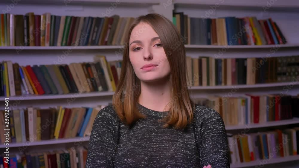 Caucasian redheaded female student watching into camera seriously and flirting on bookshelves background. Caucasian redheaded female student watching into camera seriously and flirting on bookshelves background.