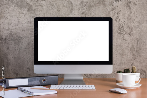 Blank screen of all in one computer with coffee cup, note book, pencil, document file and cactus vase on raw concrete background