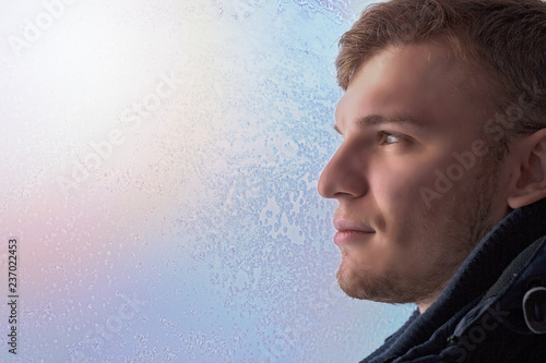 White attractive man in profile close - up against the window in the frost. Soft sunlight through the glass illuminates the person's face Winter theme. © SerPak