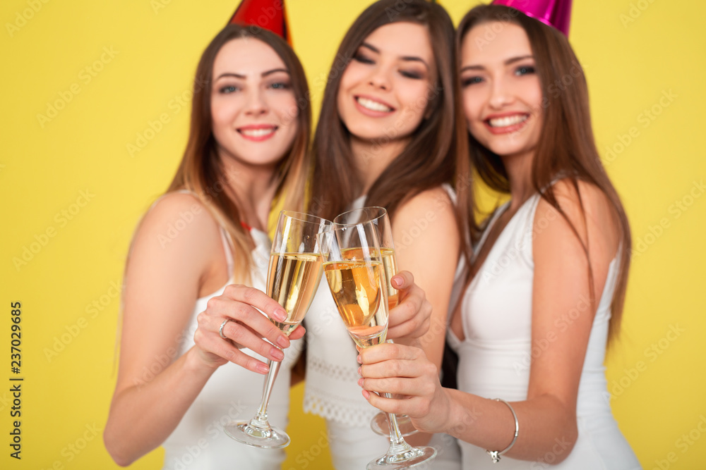 Three young woman in elegant dresses having fun, smiling, dancing and drinking champagne on yellow background. Christmas party celebration concept. Three young woman in elegant dresses having fun, smiling, dancing and drinking champagne on yellow background. Christmas party celebration concept.