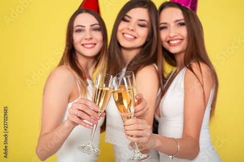 Three young woman in elegant dresses having fun, smiling, dancing and drinking champagne on yellow background. Christmas party celebration concept.